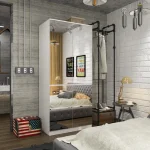 A white wardrobe with mirrored doors reflecting the bedroom interior, including a bed, bedside tables, and exposed brick walls.
